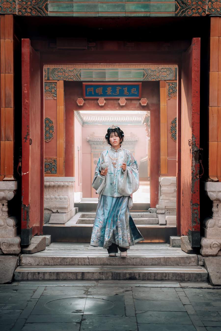Mujer con ropa tradicional china, con atuendo estilo de la dinastía Qing en tonos azules y celestes. Situada bajo el arco de un portal histórico de color rojo.