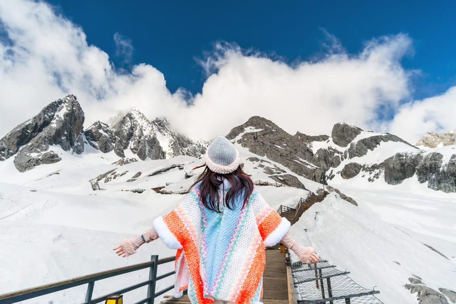 Mujer se encuentra de espaldas, con los brazos abiertos. Vestida con un poncho de colores y un gorro. Se encuentra en un pasillo de madera entre las montañas cubiertas de nieve