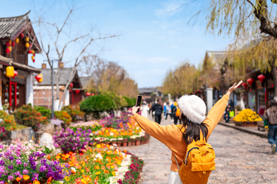 Mujer vistiendo un sueter de color naranaja, lleva puesta una mochila pequeña del mismo tono. Está en una calle peatonal de adoquines y se aprecian flores coloridas.
