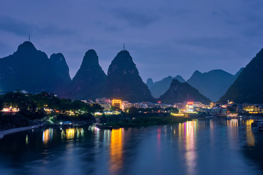 Río rodeado de un pueblo iluminado por la noche. En el fondo varias montañas
